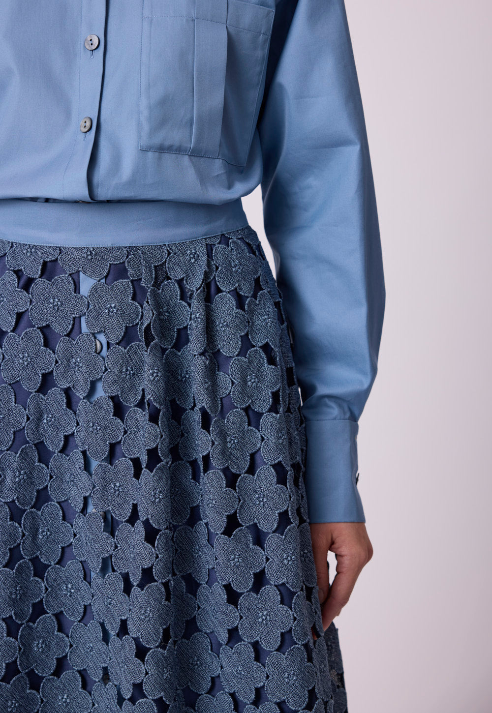 Detail view of the blue Safira blouse in crisp cotton with oversized chest pocket, tonal mother-of-pearl buttons and floral lace skirt styling.